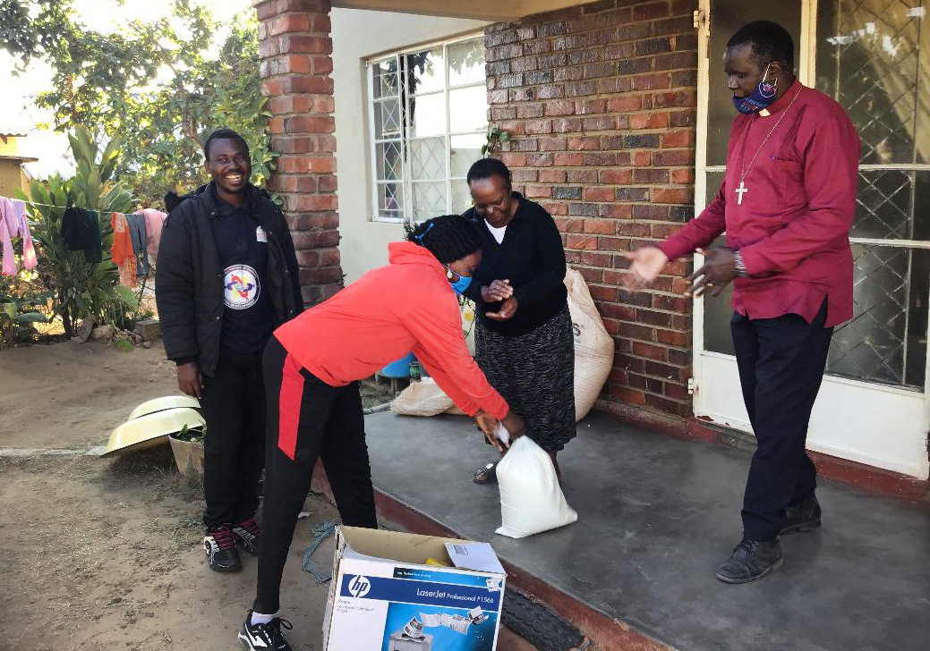 Everlyne Kukah Esther, an Africa University student from Kenya, hands over food to Charity Chikukwa, a beneficiary of the Feed a Family program in Gimboki, Zimbabwe. The Rev. Joseph Charinge (red shirt) and AU student Fiston Okito from Congo look on. Esther and her classmates started the campaign to help vulnerable families struggling during the coronavirus pandemic. Photo by Chenayi Kumuterera, UM News.