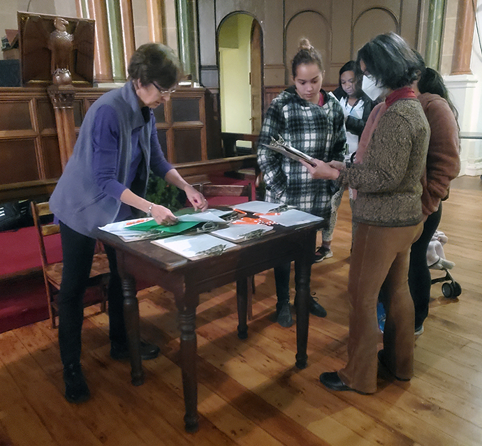 St. Paul and St. Andrew church members Karen Collins, left, and Rosangela Oliveira, right, offer forms to asylum seekers to complete so they can register for services provided by the West Side Campaign Against Hunger. Photo by K Karpen.
