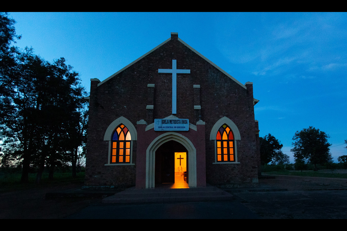 A luz se derrama da porta e dos vitrais da Igreja Metodista Unida Central de Quéssua, parte da Missão Quéssua no leste de Angola. Estabelecida no final de 1800 por missionários Metodistas e destruída por quase três décadas de guerra civil que terminou em 2002, a missão está sendo reconstruída, levando educação, saúde e segurança alimentar às pessoas no vale rural perto da capital provincial de Malanje. Foto de Mike DuBose, Notícias da MU.