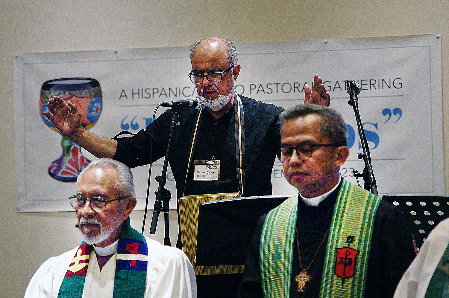 O Rev. Tercio Junker (centro), pastor da Conferência Anual do Norte de Illinois, conduz a oração para consagrar os elementos do sacramento da comunhão. Abaixo à esquerda, Dom Elías Galván, atualmente aposentado e fundador da Conferência Anual do Desierto del Suroeste atualmente liderada por Dom Carlo Rapanut à direita da imagem.