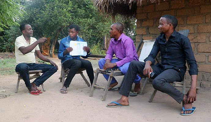 Michel Tahindja Tashinga (da esquerda) e Kitete Kahodi, estafetas da comunidade Metodista Unida baseados nas aldeias de Wenga e Dikungu, respetivamente, falam com Patrice Lumumba Okeke (de purpura) e outro residente sobre a varíola. "Graças à informação dada, posso reconhecer os sintomas e saber o que fazer se os notar em mim ou noutra pessoa da minha família," disse Okeke, que lidera uma família de oito pessoas. Foto de Chadrack Tambwe Londe, Notícias da MU.