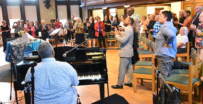 The audience holds hands and sings "Draw the Circle Wide in the chapel of Drew Theological School to commemorate the opening of the new LGBTQ+ United Methodist Heritage Center in Madison, N.J. Musician Mark Miller played the piano and led the audience in song. Photo by John Coleman, UM News.