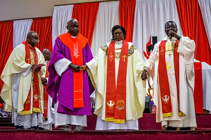 Les évêques Katembo (à droite, en violet) et Kasap Owan (à gauche, en blanc) tiennent les mains de l'évêque Mujinga Kashala (au centre) lors de la cérémonie d'installation de cette dernière à Lubumbashi, en République démocratique du Congo. L'évêque Mande Muyombo est visible à l'arrière-plan, à l'extrême gauche. La cérémonie a rassemblé des centaines de fidèles, des dirigeants religieux (dont le Révérend André Bokundoa et plusieurs évêques émérites) ainsi que des autorités politiques locales et nationales. Photo avec l’aimable autorisation du Département de Communications de la Région Épiscopale du Sud-Congo et la Zambie.